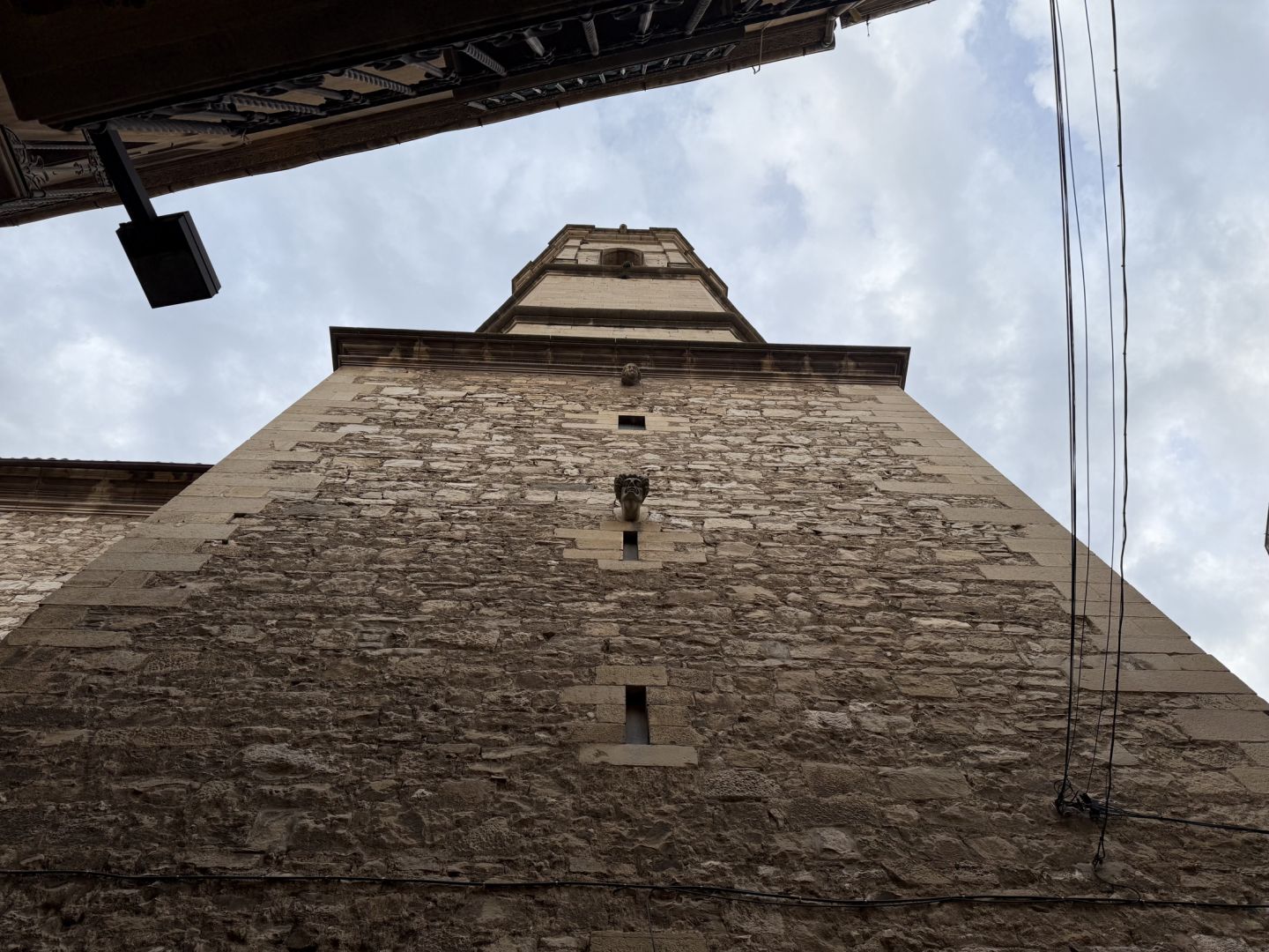 El campanar de Tàrrega, vist des de baix, centrat dins l’àrea de la imatge i ocupant-la gairebé tota; la vista és des de baix en punt de fuga cap amunt; a la part superior s’hi veu un fragment de la façana de l’edifici del davant.