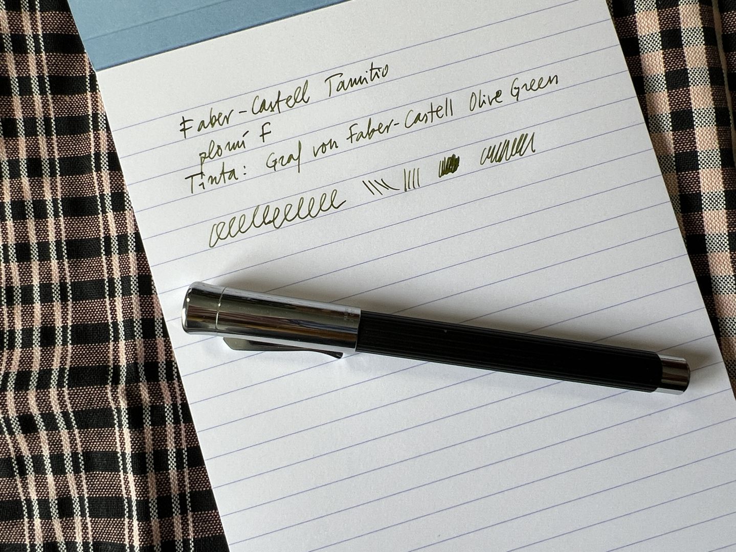 Full d’un bloc de mida A5 amb pauta. Text: Faber-Castell Tamitio, plomí F, tinta Graf von Faber-Castell Olive Green. A sota, la ploma en qüestió. Fons: un drap de roba de fer farcells.