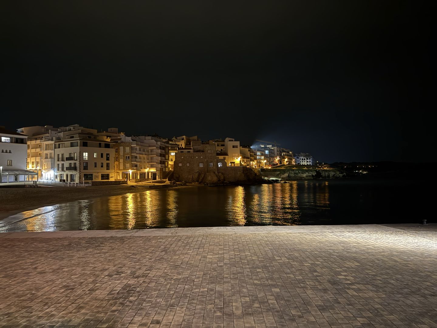 Vista d’una part de l’Escala des d’un extrem de la cala cèntrica. Filera d’edificis horitzontal al centre, amb llums blanques i grogues dels diferents carrers i locals reflectint-se a l’aigua del mar. Cel negre del tot.